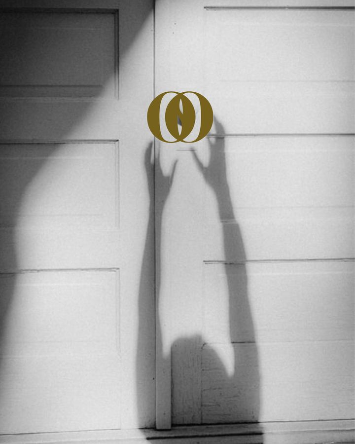 Zwart-witfoto van een schaduw van een reikende hand onder het gouden She Society-logo op een witte deur