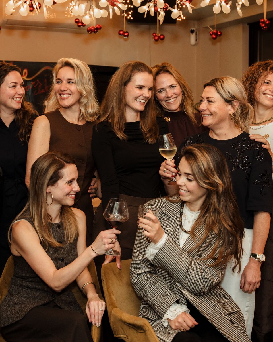 Negen vrouwen lachen en proosten samen tijdens een She Society-avond, onder feestelijke verlichting
