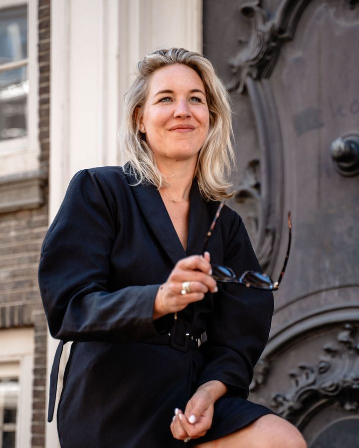 Portret van Robin, oprichter van She Society, lachend in een donkere blazerjurk met een zonnebril in de hand